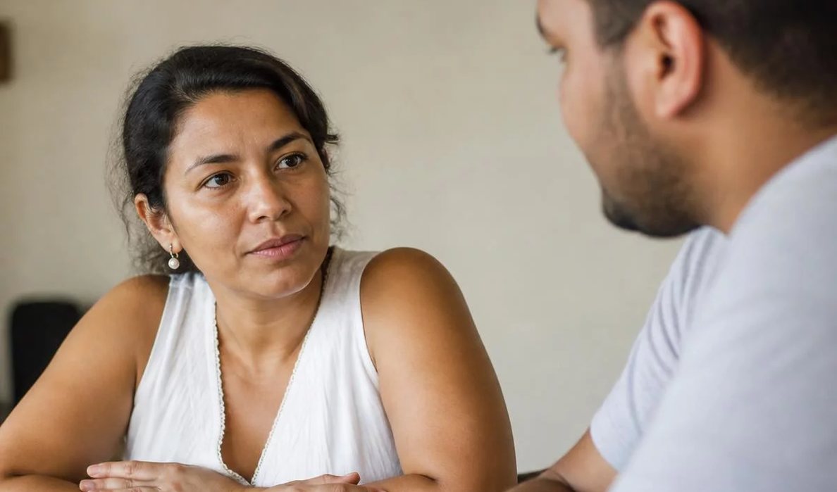 Três participantes conversam ao redor de uma mesa comprida numa sala clara de janelas altas